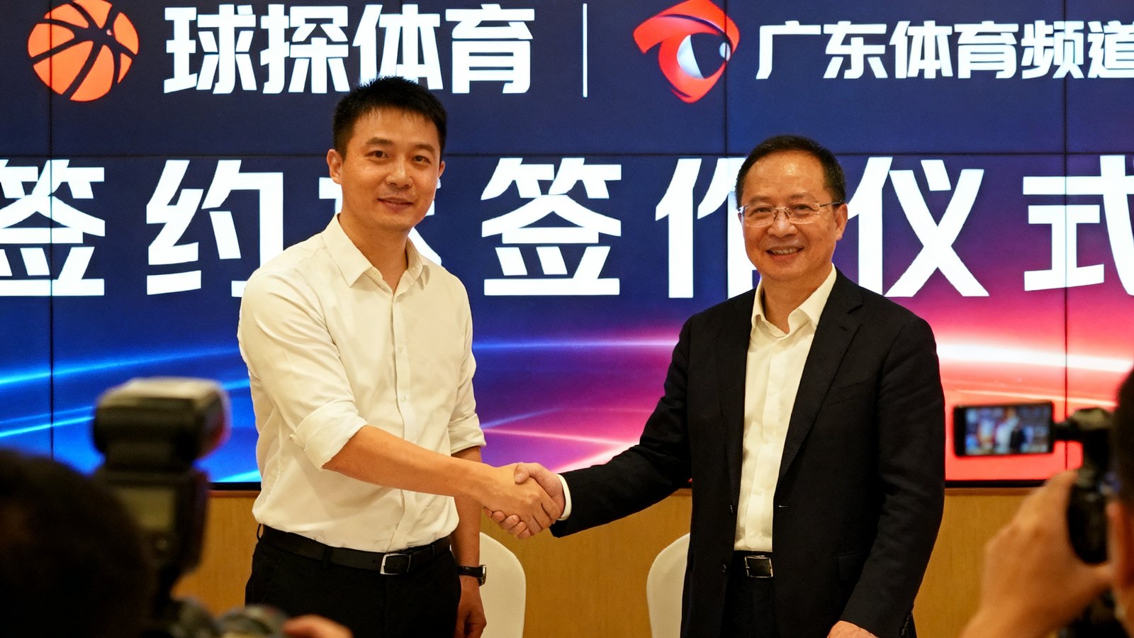 签约仪式现场照片，球探体育创始人张明与广东体育频道总监在签约台前握手，背景展示板醒目显示双方品牌标识，现场有媒体记者拍摄记录这一合作时刻。